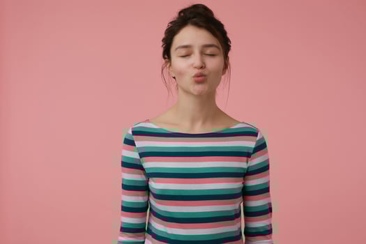 Portrait of attractive, lovely girl with brunette hair and bun. Wearing striped blouse and try to kiss with eyes closed. Emotional concept. Stand isolated over pastel pink background photo