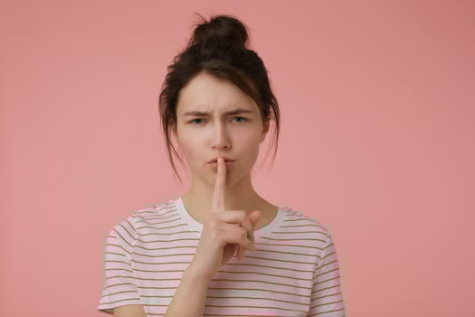Teenage girl, worried woman with brunette hair and bun. Wearing t-shirt with red strips and showing sign of silence. Emotional concept. Watching at the camera isolated over pastel pink background photo