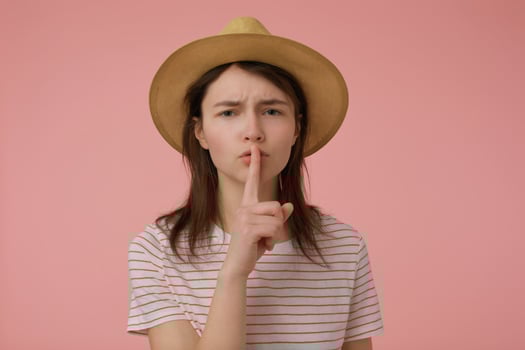 Teenage girl, woman with long brunette hair frown. Wearing t-shirt with red strips and hat. Show silence sign. Emotional concept. Watching at the camera isolated over pastel pink background photo