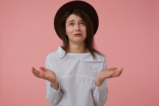 Upset looking woman, unhappy girl with long brunette hair. Wearing white blouse and black hat. Emotional concept. Stand isolated over pastel pink background and watching up at copy space photo