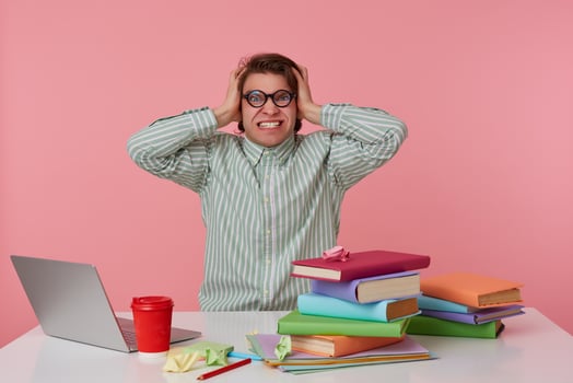 Young angry student in glasses, sits by the table and working with laptop, holds his head and looks surprised , isolated over pink background. photo