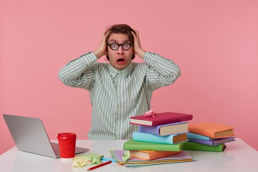 Portrait of young angry man in glasses, sits by the table and working with laptop, holds his hesd and looks surprised , isolated over pink background. photo