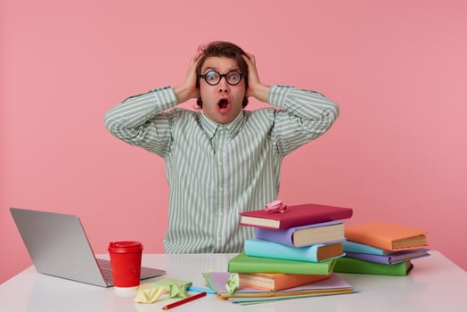 Young surprised student in glasses, sits by the table and working with laptop, covers his ears and shouts , isolated over pink background. photo