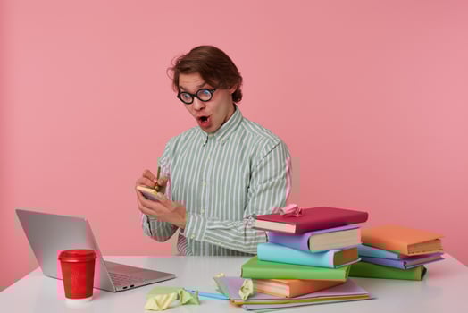 Shocked young man in glasses sits by the table and working with laptop, writes the solution of the equation on the stickers isolated over pink background. photo