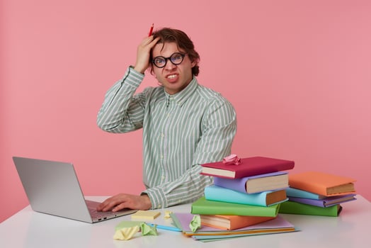 Portrait of angry young guy in glasses sits by the table and working with laptop, very dissatisfied, holds hand on forehead with wide open mouth, isolated over pink background. photo