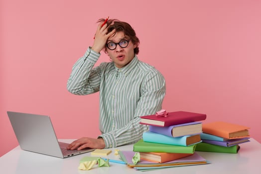 Photo of unhappy young man in glasses sits by the table and working with laptop, very dissatisfied, holds hand on forehead isolated over pink background.