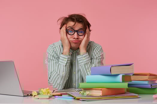 Studio photo of young male with wild hair leaning head on hand hands and keeping palms on face, felling tired after long busy day, posing over pink background at working table