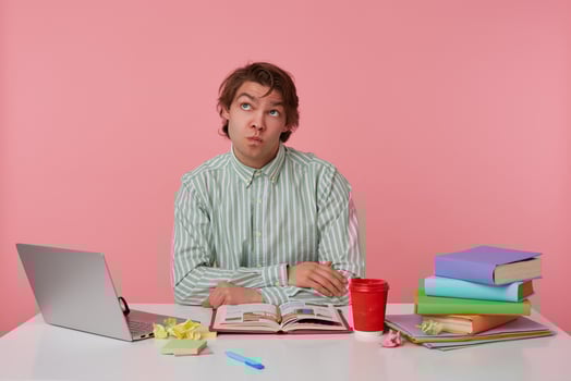 Indoor shot of thoughtful young dark haired male posing over pink background, sitting at working table with folded arms on it, looking upwards with pursed lips and raising eyebrows photo