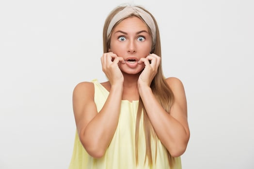 Shocked young long haired blonde female in beige headband raising scaredly hands to her face and rounding her blue eyes while looking amazedly at camera, isolated over white background photo