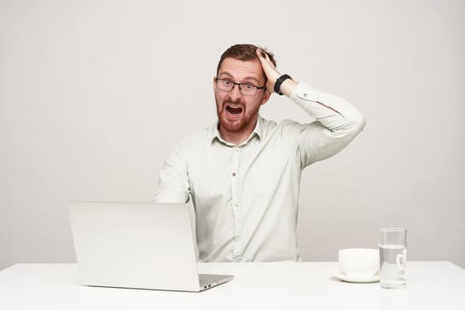 Indoor photo of young agitated bearded male dressed in white shirt clutching his head with raised hand and looking amazedly at camera, posing over white background