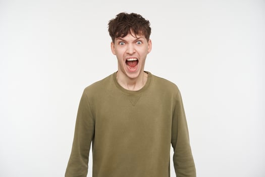 Young man, angry guy with brunette hair, piercing and bristle. Wearing khaki color sweater. Yelling hard, shout in anger. Watching at the camera isolated over white background photo