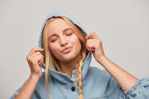 Attractive beautiful blonde young woman with braid, hands keep hood, sends air kiss with lips, eyes closed, feels love, wears oversize jeans denim coat over white background photo