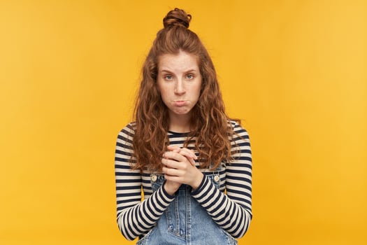 indoor portrait of young displeased ginger girls, keeps her palms together, curves her lips with a sad facial expression. isolated over yellow background. photo