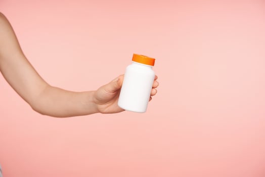 Studio shot of lady's hand holding white bottle with orange cover while posing over pink background, going to take vitamin with glass of water. Healthcare concept photo