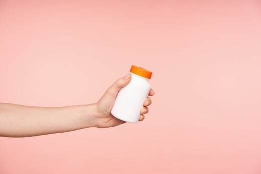 Studio photo of young female's hand with nude manicure keeping bottle with pills in it while being isolated over pink background. Healthcare and beauty concept