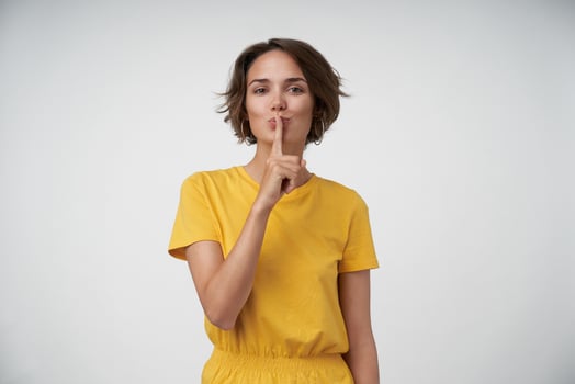Attractive young female lady with short brown hair keeping fogefinger on her lips, showing hush gesture, asking to keep secret, posing over white background in casual clothes photo