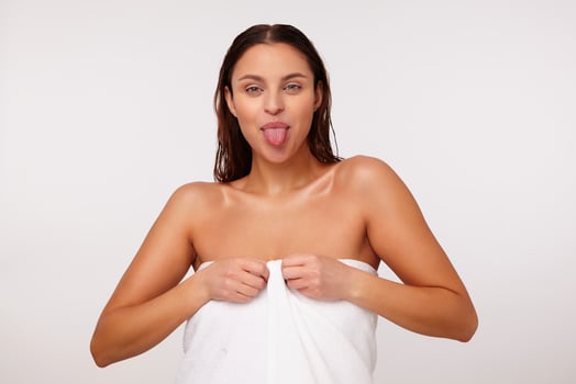 Indoor photo of young beautiful cheerful dark haired female showing tongue while looking positively at camera, standing over white background in bath towel