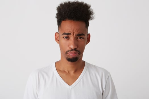 Studio photo of young offended dark skinned brunette man with trendy haircut pouting his lips while looking sadly at camera, isolated over white background
