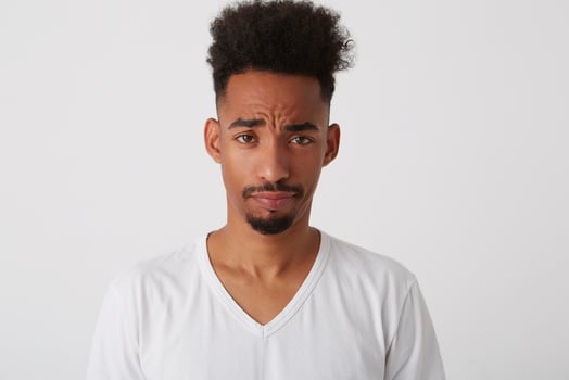 Close-up of young attractive dark skinned bearded guy frowning his eyebrows while looking at camera while standing over white background in basic t-shirt photo