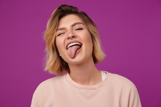Indoor portrait of young joyful funny dyed blonde female, with tattooed arms showing her tongue, isolated over purple background photo