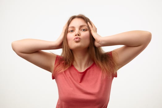 Studio photo of young blonde lady with loose long hair folding her lips in air kiss and holding head with raised hands while standing over white background