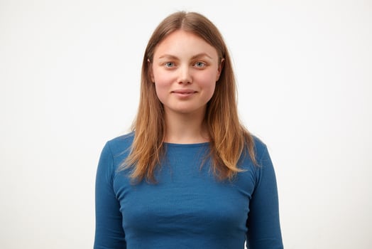 Positive young pretty blonde woman with natural makeup smiling gently while looking at camera, keeping hands down while standing over white background photo