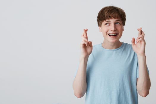 Teenager looks away, wishing something pleasant, hands up and fingers twisted, with braces on teeth, wears blue t-shirt,blank copy space for your promotional content isolated over white background photo