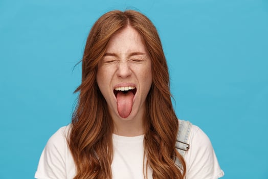 Funny shot ot young joyful redhead lady with natural makeup fooling while posing over blue background, frowning face and sticking out her tongue with closed eyes photo
