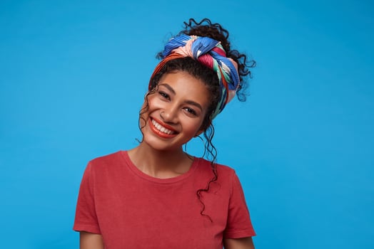 Happy young cute brunette curly female with nose piercing showing her pleasant emotions and smiling cheerfully while looking at camera, isolated over blue background photo