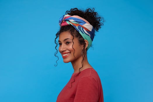 Side view of young cheerful brunette curly woman with festive makeup smiling broadly while looking gladly ahead, isolated over blue background in casual clothes photo