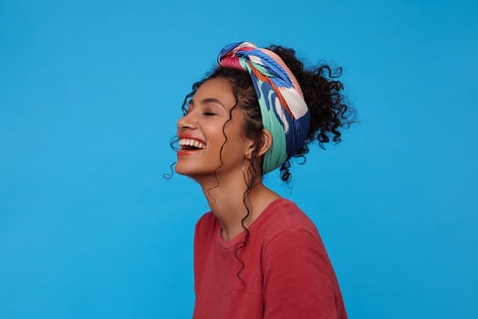 Pleasant looking young cheerful dark haired curly lady throwing back her head while laughing happily with closed eyes, standing over blue background with hands down photo