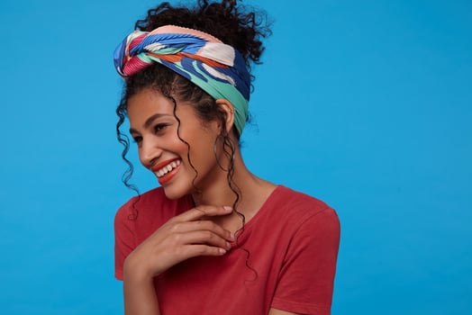 Happy young lovely brown haired curly female with gathered hair smiling happily and touching her neck with raised hand, isolated over blue background photo