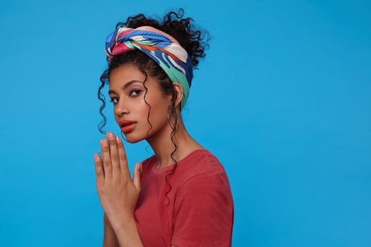 Serious young pretty brunette woman with casual hairstyle keeping raised palms together while looking pensively at camera, isolated over blue background photo