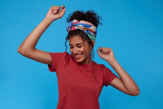 Good looking young pretty brunette curly woman with gathered hair smiling cheerfully with closed eyes while dancing with raised hands, posing over blue background photo