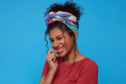 Coquettish young lovely dark haired curly woman giving gladly wink at camera and keeping finger on underlip while looking cheerfully at camera, isolated over blue background photo