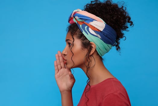 Side view of young attractive dark haired curly female with gathered hair keeping raised palm near her mouth while going to tell something secret, isolated over blue background photo