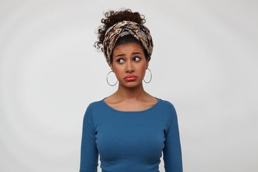 Offended young lovely brunette dark skinned female twisting her mouth while looking sadly aside, keeping hands along body while standing over white background photo