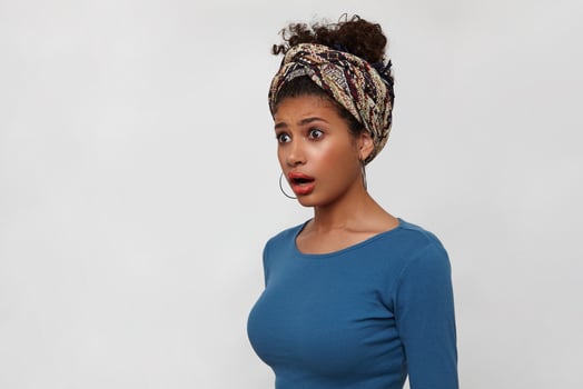 Bemused young dark haired curly female with gathered hair looking dazedly aside with wide eyes and mouth opened, standing over white background with hands down photo