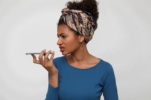 Displeased young curly brunette dark skinned female grimacing her face while having unpleasant phone conversation, standing over white background with smartphone photo