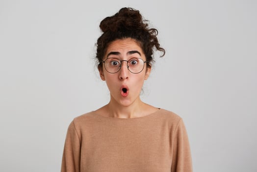 Amazed shocked young woman with bun of dark curly hair wears beige pullover and glasses looks surprised and impressed isolated over white background Looking directly in camera photo