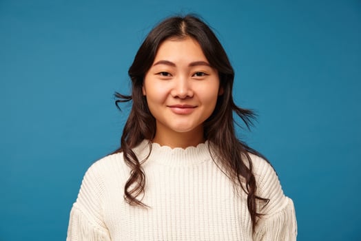 Indoor photo of pleasant looking young dark haired asian female with wavy hairstyle smiling positively at camera while standing against blue background