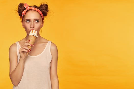 Young lady, pretty ginger woman with two buns. Wearing white tank top and red doted hairband. Eating an ice cream. Watching to the right at copy space, isolated over yellow background photo