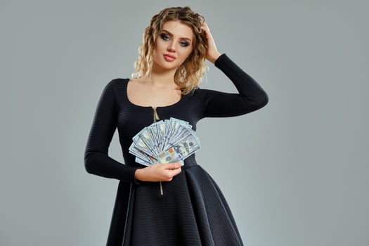 Blonde female in black short dress is holding some cash, posing against gray background. Gambling entertainment, poker, casino. Close-up. photo