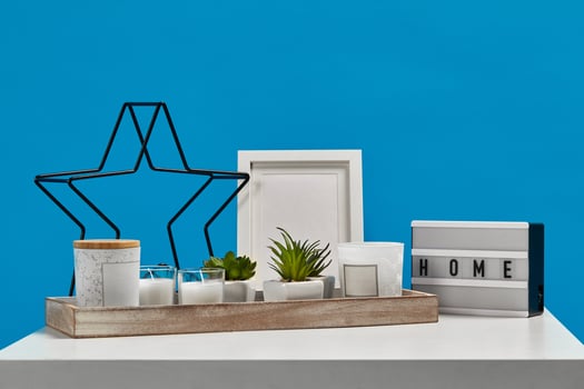 White table, wooden stand with green succulents, candles, jars. Black iron star, lamp inscription home, empty photo frame. Blue background. Close up
