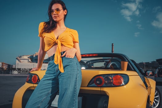 Girl in blue jeans, orange top and sunglasses is posing leaning on yellow car cabriolet with paper cup of beverage on trunk at parking lot. Copy space photo