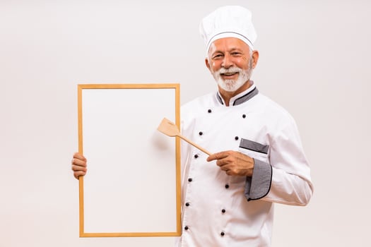 Portrait of senior chef showing with wooden spoon whiteboard on gray background. photo