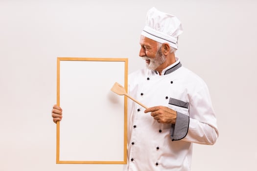 Portrait of senior chef showing with wooden spoon whiteboard on gray background. photo