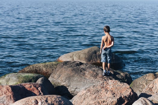 A little lonely boy stands on large stones and looks at the sea. photo