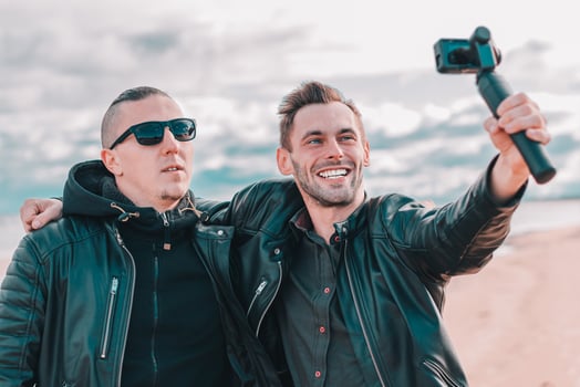 Two Handsome Smiling Friends Making Selfie Using Action Camera with Gimbal Stabilizer at the Beach. Youthful Men in Black Clothes Having Fun by Making Photos