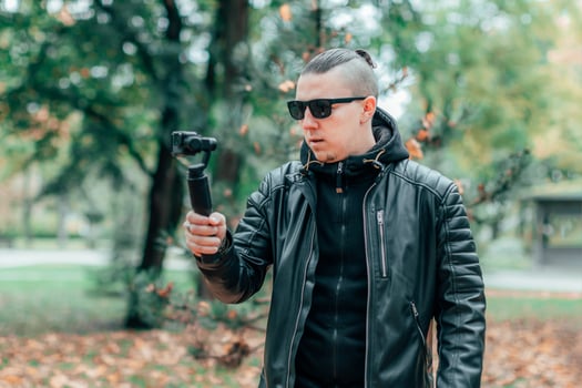 Blogger in Black Clothes and Sunglasses Making Video Using Action Camera with Gimbal Camera Stabilizer in Autumn Park photo